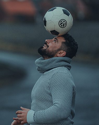 A man in grey sweater holding a soccer ball on his head.